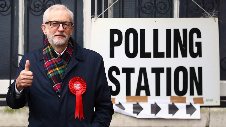 A man wearing a scarf does a thumbs up next to a sign that reads polling station