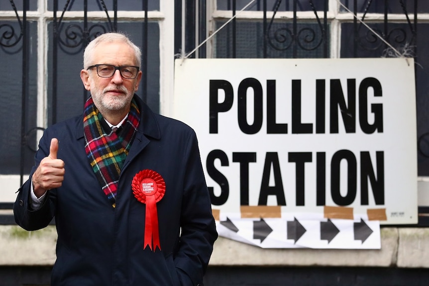 A man wearing a scarf does a thumbs up next to a sign that reads polling station