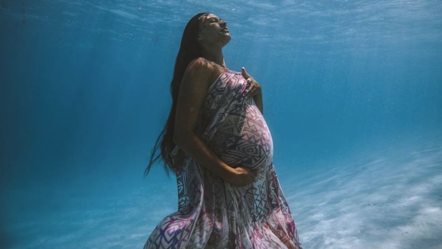 Heavily pregnant woman stands underwater draped in ie lavalava, eyes closed her face is held up towards sunrays on sea'surface. 
