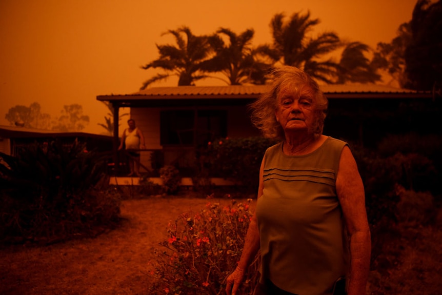 An older woman stands with worried and bewildered expression in front of home, a the entire scene a dark orange hue.