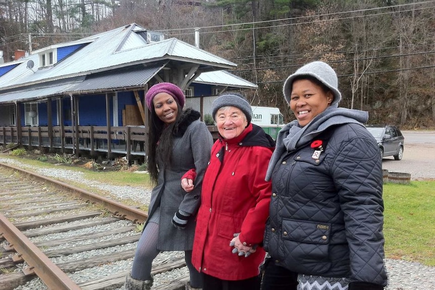 Three smiling women in winter clothes link arms while standing on a train track.