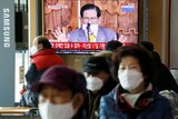 A man speaks on a TV screen in front of people wearing masks.