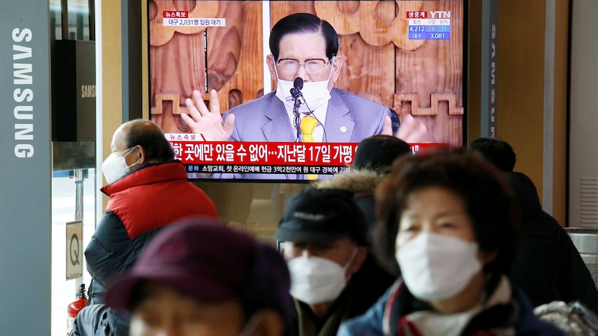 A man speaks on a TV screen in front of people wearing masks.