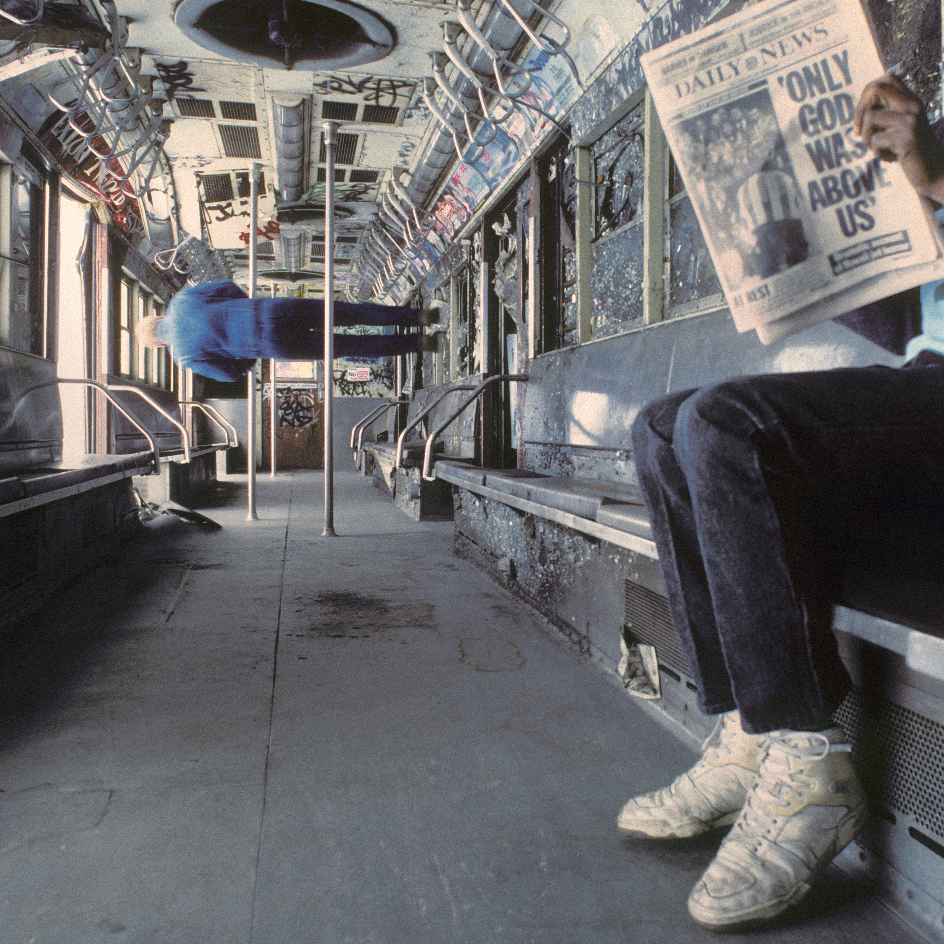 an empty train car with one man appearing to stand on one of the walls. someone sits and reads a newspaper.