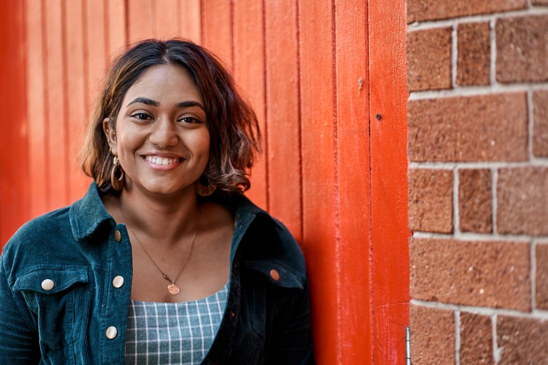 A young South Asian woman smiles standing against a red wall.