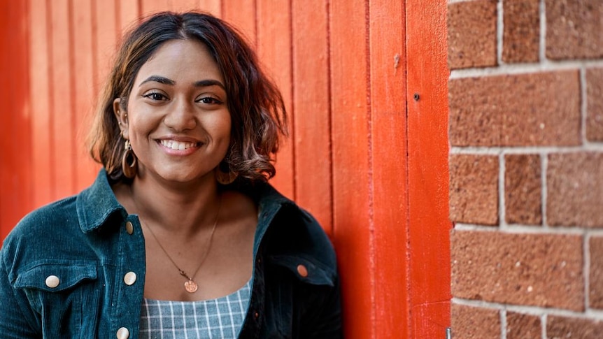 A young South Asian woman smiles standing against a red wall.