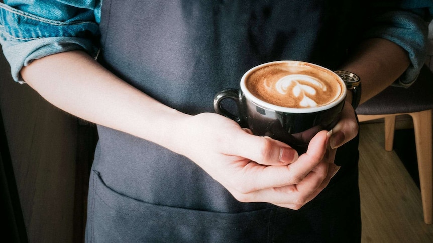 A close-up shot of a barista, wearing a dark apron and holding a coffee in both hands.