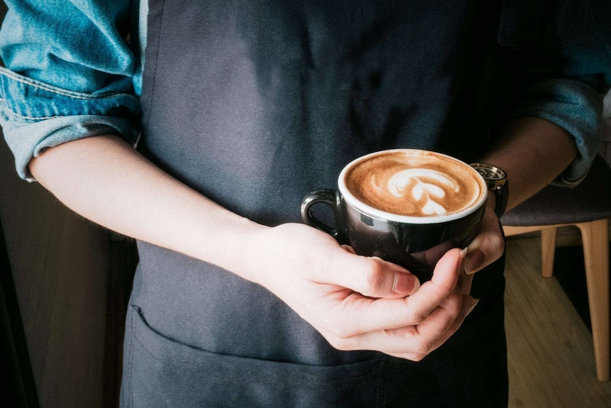 A close-up shot of a barista, wearing a dark apron and holding a coffee in both hands.