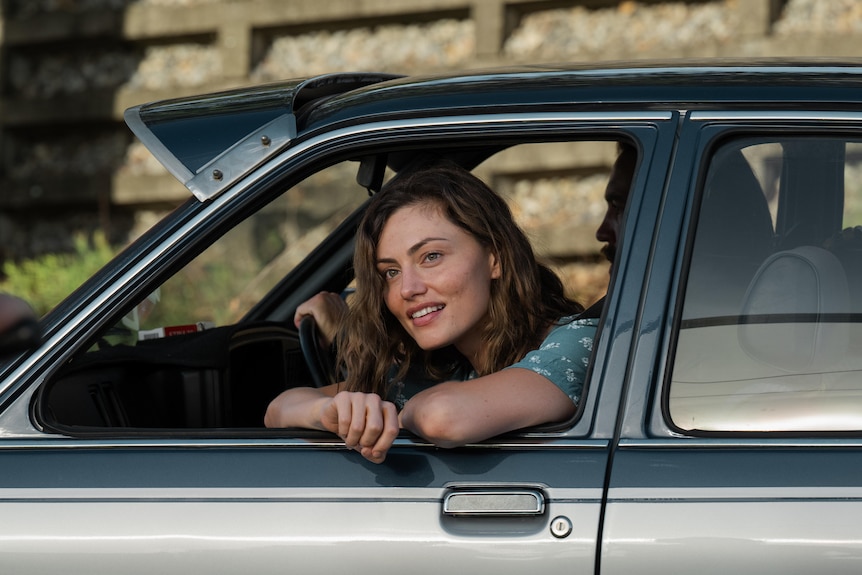 Phoebe, as Frances, leans out a car window and smiles while looking into the distance.