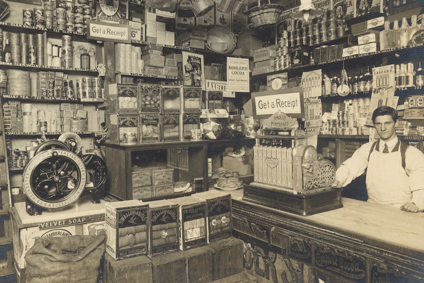 A sepia photo of a grocery store in the early 1900s - with shelves full of products and a man with an old cashier