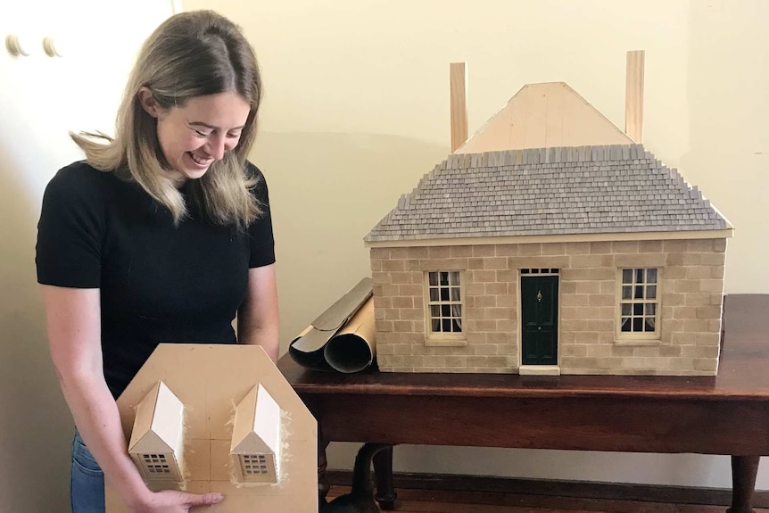 Effie Pryer and her cat puss in front of the dolls house version of a colonial sandstone cottage,