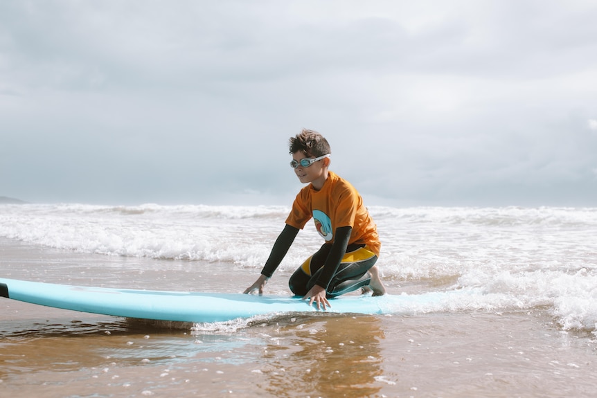 A young boy wearing an orange shirt and goggles rides a wave close to the shore.