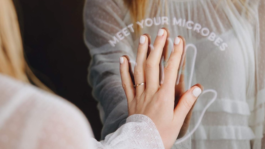 A woman with long blonde hair puts her hand on the outline of a hand on a mirror with 'Meet Your Microbes' written on it.