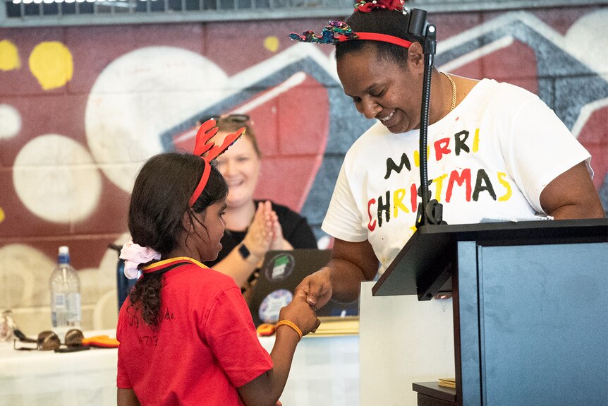 A student in a red shirt wearing reindeer antlers shaking hands with a teacher wearing a shirt that says Murri Christmas.
