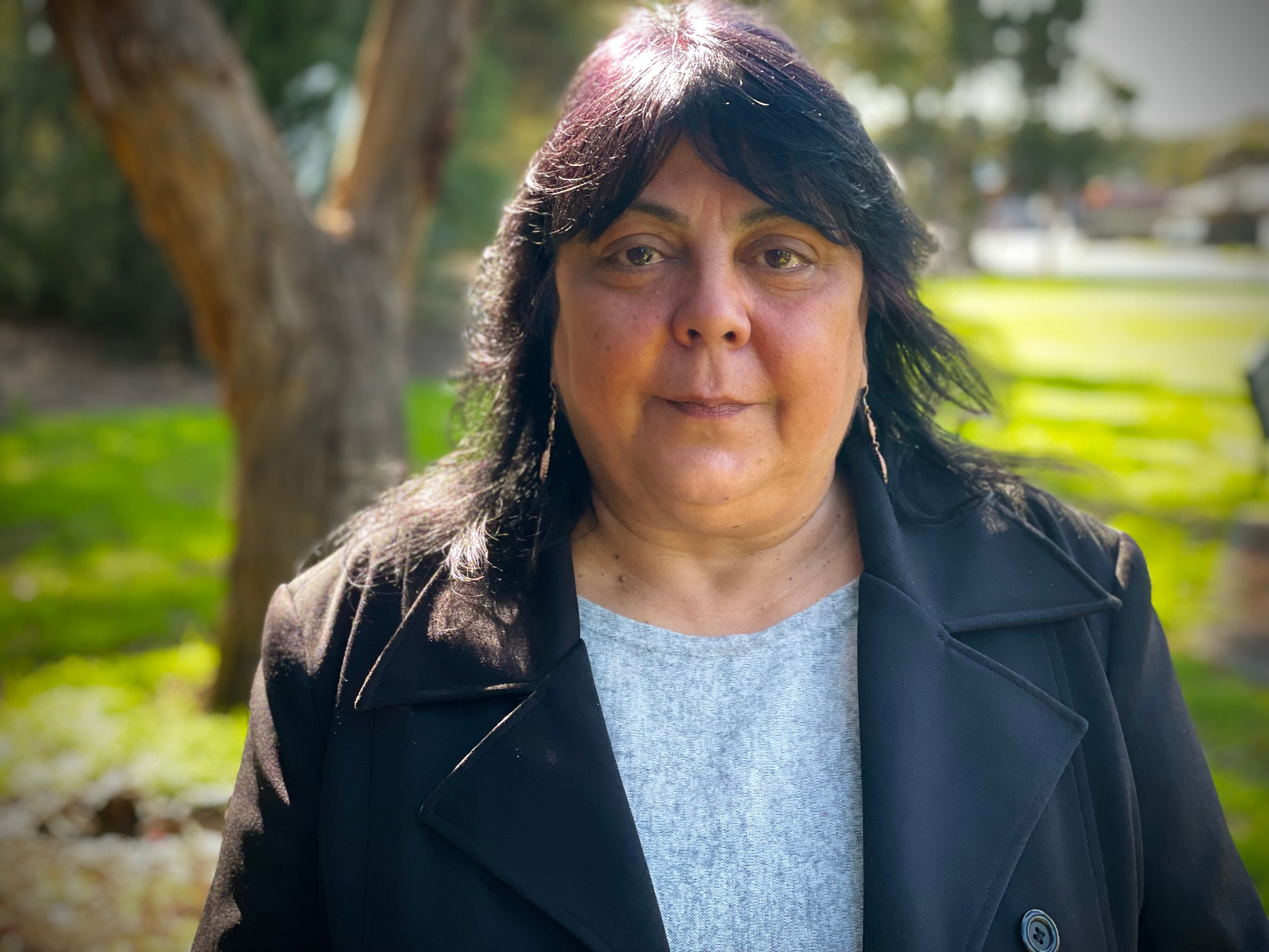 A portrait of Cheryl Axleby looking into the camera, as she stands in what looks like a leafy green park.