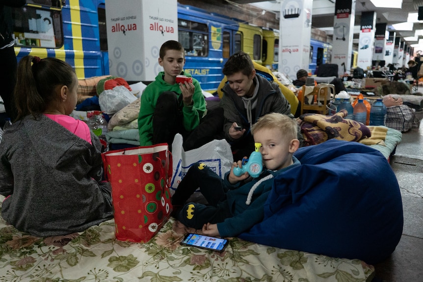 A kid shows off his little blue plastic toy while sitting on a train platform with a group of kids