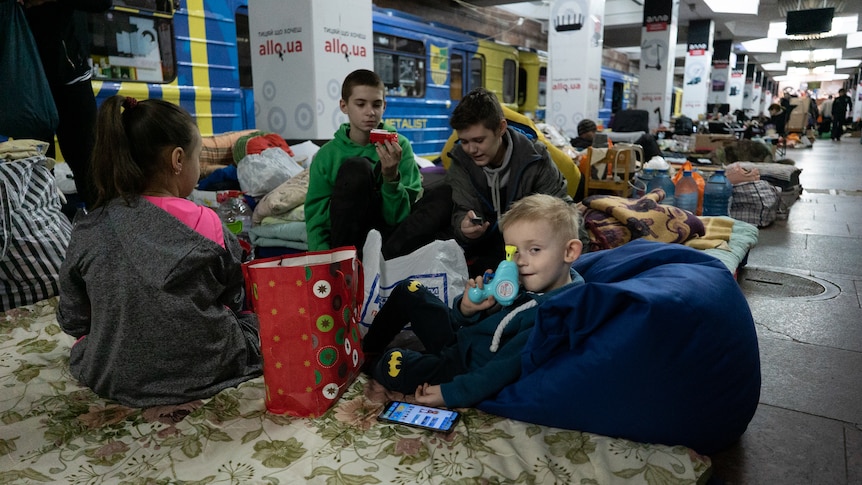 A kid shows off his little blue plastic toy while sitting on a train platform with a group of kids