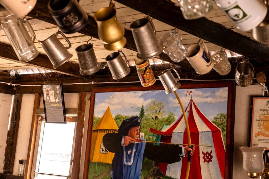 Large beer cups hang from a ceiling with a painting of a archer in medieval clothing hangs on the wall.