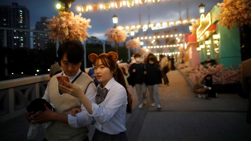 A South Korean woman wearing bear ears hold up her phone for her boyfriend to look at.