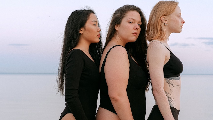 Three women, of different sizes, wearing black and standing on beach.