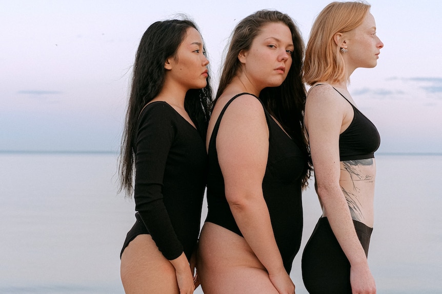 Three women, of different sizes, wearing black and standing on beach.
