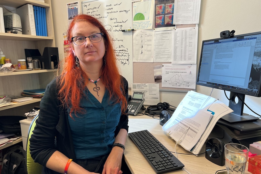 Someone with red hair and glasses sits at a desk.