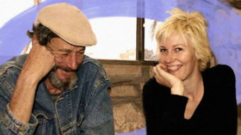 A man smiles while looking down. A young woman sits next to him smiling towards the camera.