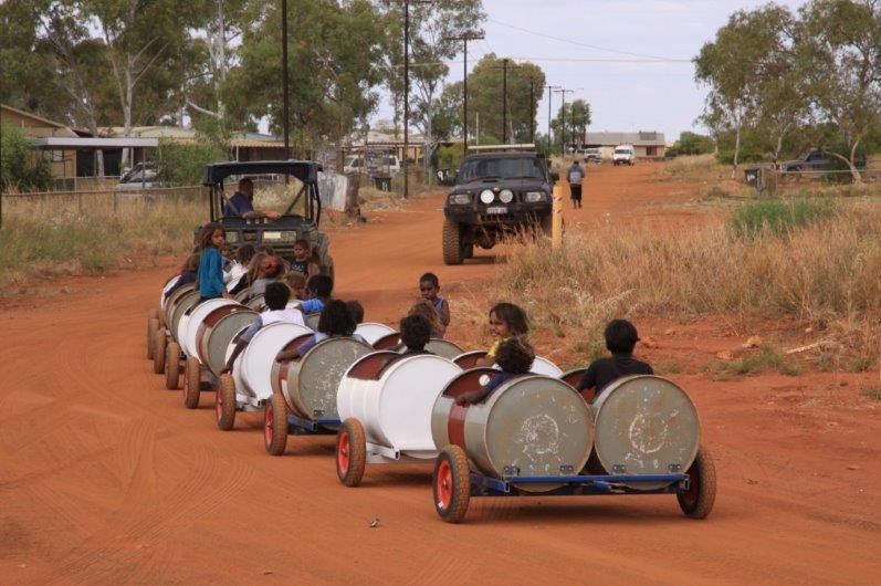 Kids ride in barrels being pulled behind a tractor though an outback community.