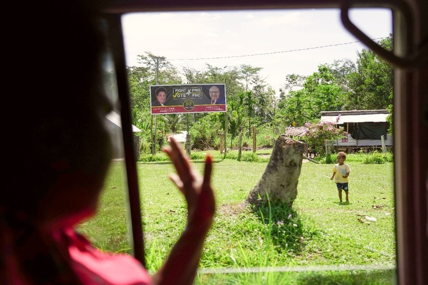 A woman waves through a car window towards a child standing in a field in front of an election sign