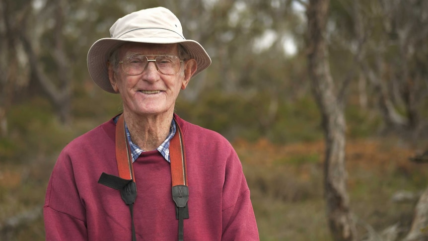 An elderly man smiling with a hat on, a red jumper and a pair of binoculars hanging round his neck.