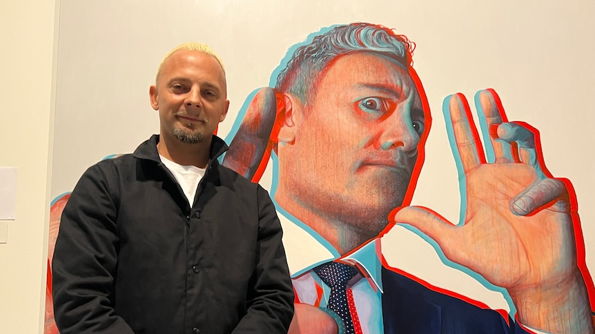 White man with blonde hair and black coat stands with portrait of Taika Waititi, a grey-haired man in a navy suit in 3D style