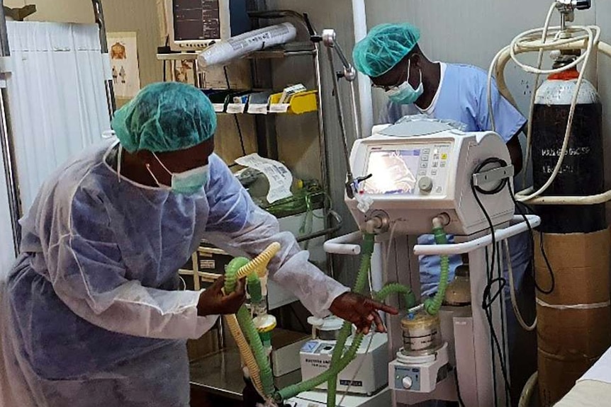 Two medical staff in protective clothing, masks and green caps check medical equipment.
