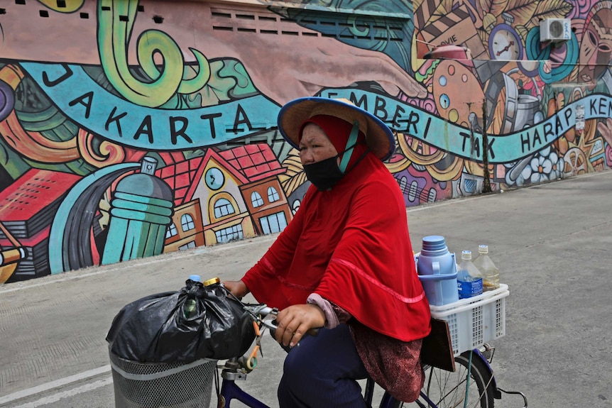 A woman selling a traditional herbal drink rides a bike and wears a mask past a mural in Jakarta.