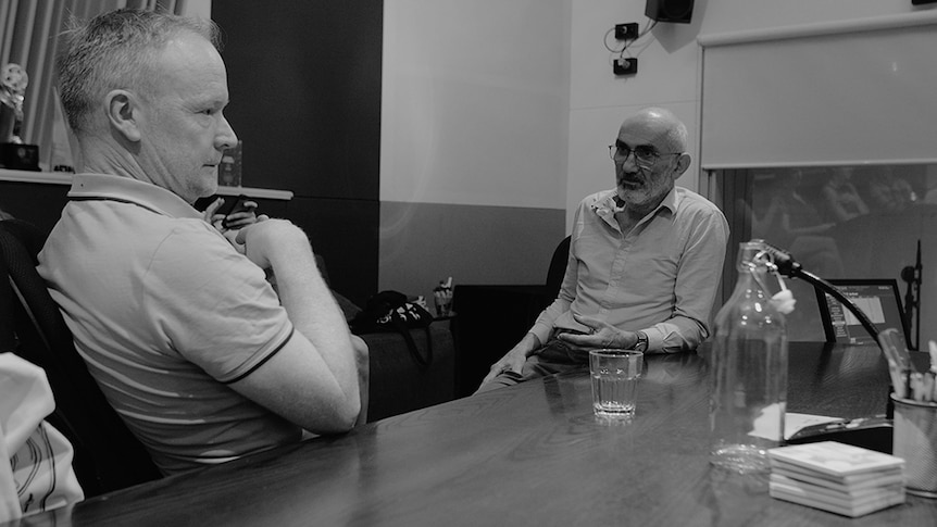 A black and white photos of James Ledger and Paul Kelly sitting at a table facing each other.