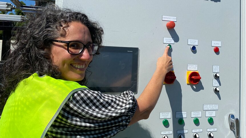 A smiling woman with curly hair and glasses hits a button on a grey metal pyrolysis technology machine with colourful buttons.