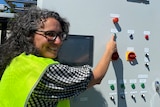 A smiling woman with curly hair and glasses hits a button on a grey metal pyrolysis technology machine with colourful buttons.