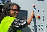 A smiling woman with curly hair and glasses hits a button on a grey metal pyrolysis technology machine with colourful buttons.