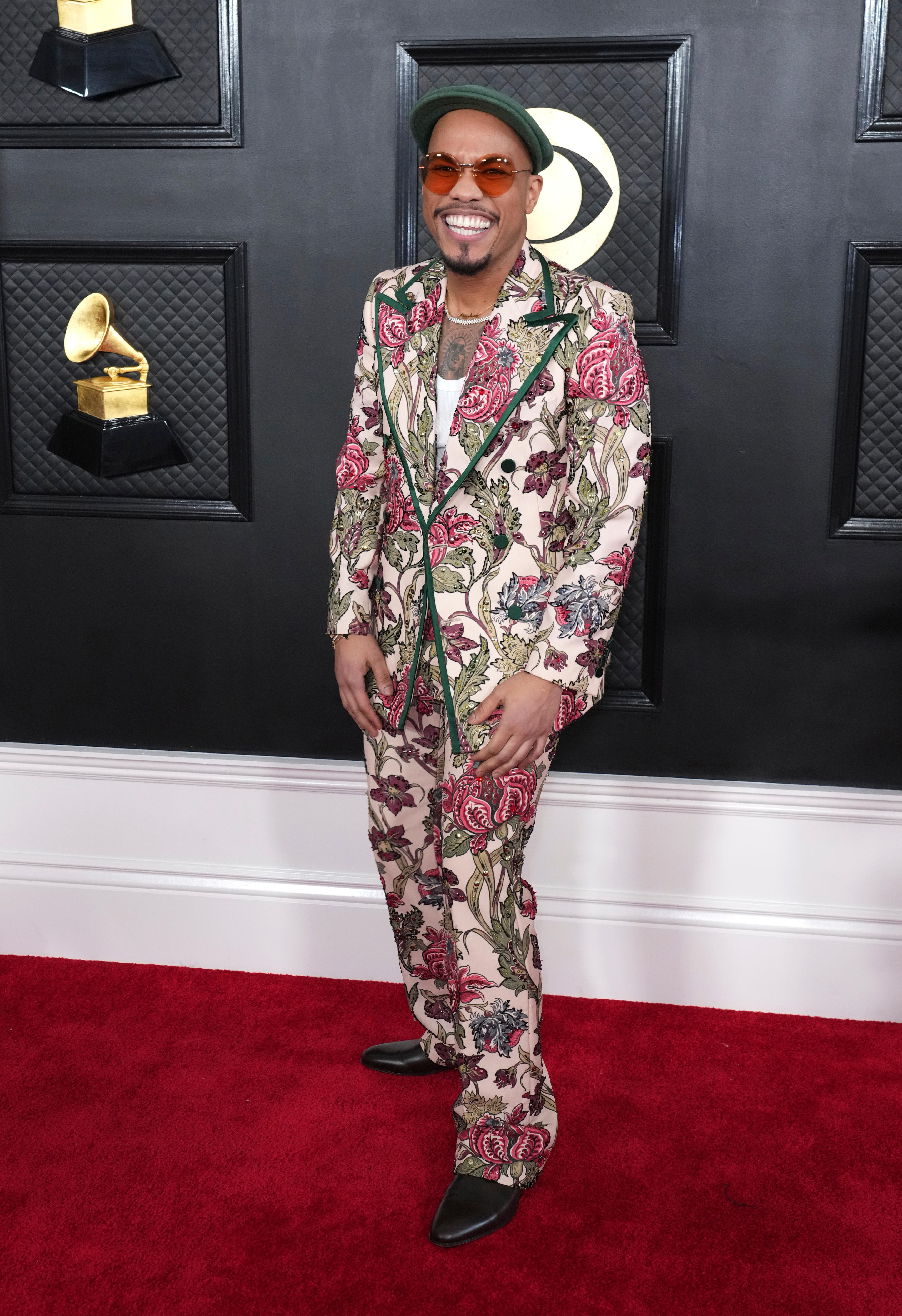 Anderson .Paak wearing a dark pink and green floral suit with yellow glasses and a dark green cap. 