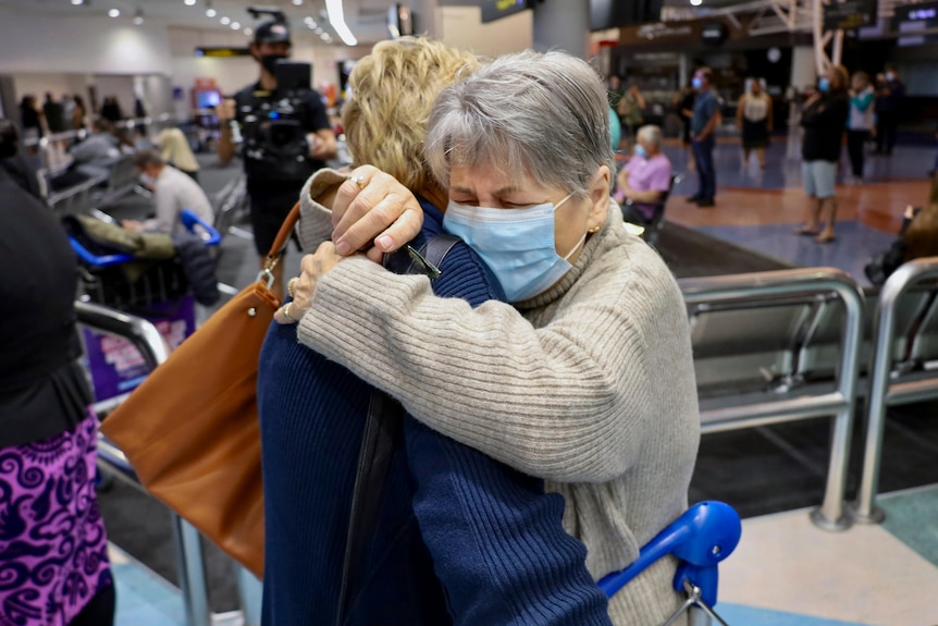 Woman with short grey hair wearing a mask hugs another woman and looks overcome with emotion.