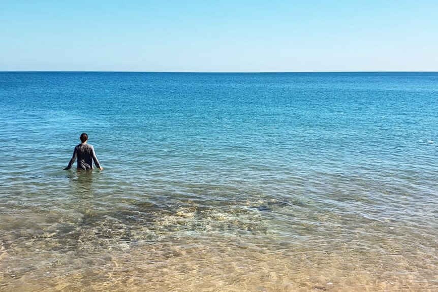 A woman stands in the water of a flat sea.