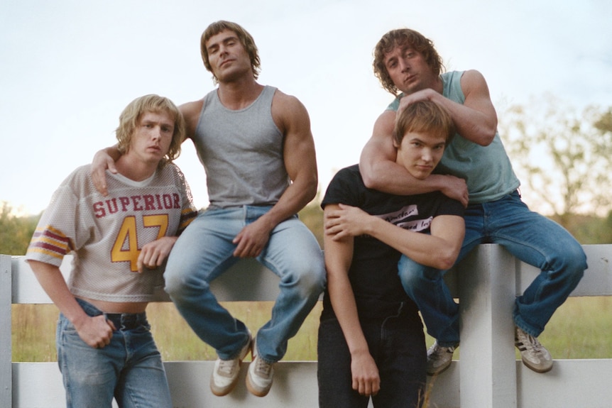 Four beefed up brothers in jeans and singlets or t-shirts, sit or lean together on a farm fence.