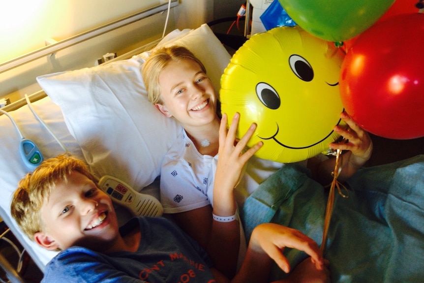 Two young children with blonde hair lie in a hospital bed smiling at the camera holding balloons 