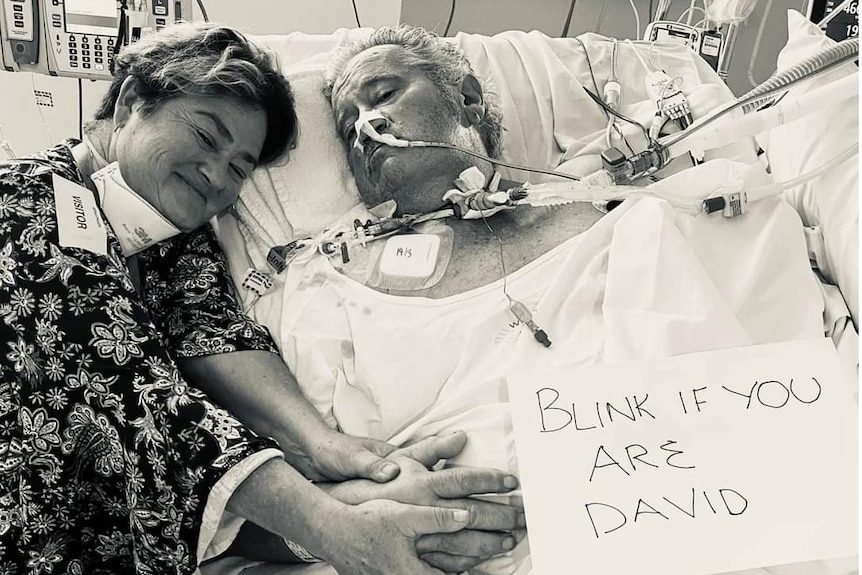 A woman holds hands with a man lying a hospital bed, who has a sign saying 'Blink if you are David'