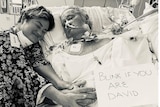 A woman holds hands with a man lying a hospital bed, who has a sign saying 'Blink if you are David'