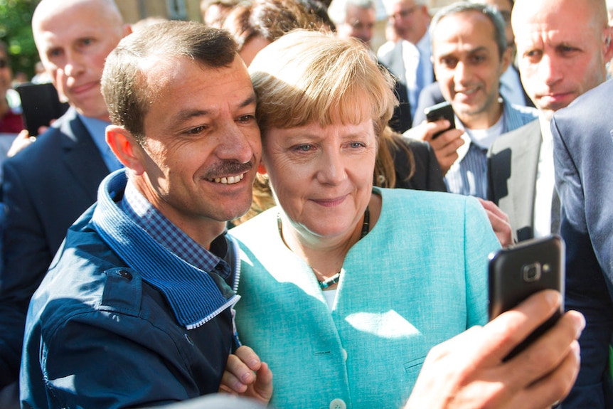 German chancellor Angela Merkel standing next to a man as he holds up his mobile phone and takes a photo of them together