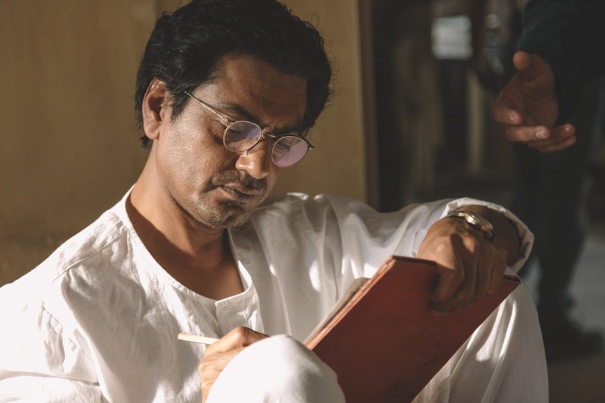 An Indian man in white clothing sits on the floor against a wall, writing on a piece of paper.