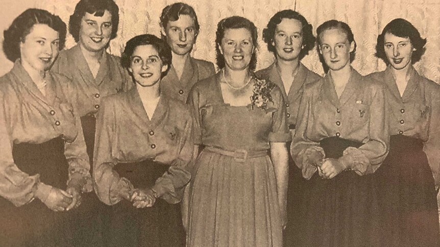 Group of 1950s teenage girls in long dresses posing for photograph with their boss, a middle aged woman