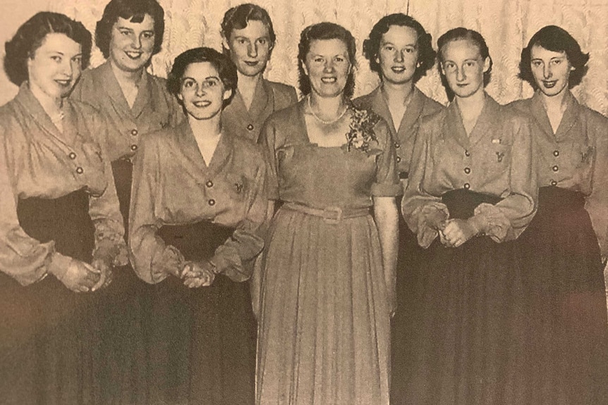 Group of 1950s teenage girls in long dresses posing for photograph with their boss, a middle aged woman
