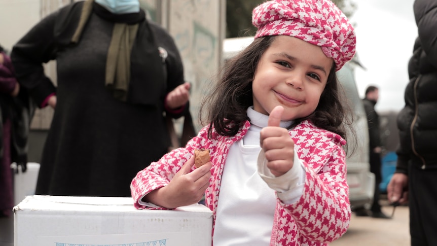 A very young girl in a pink and white outfit gives a thumbs-up while leaning on a food parcel