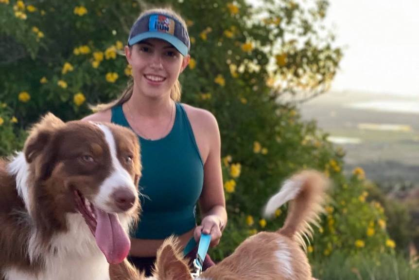 A young woman smiles as she stands on a hilltop holding two dogs on leads.